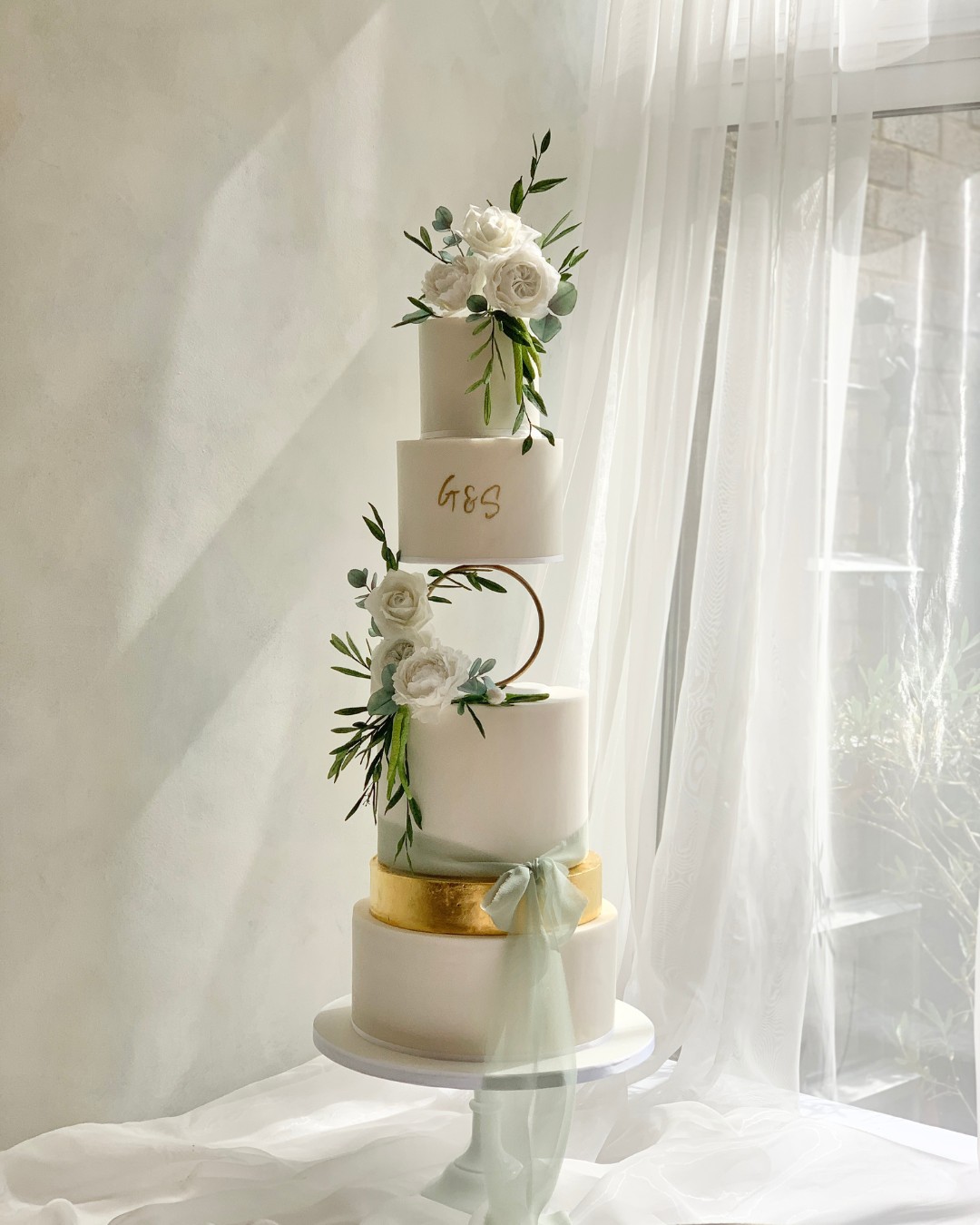 large 5 tier wedding cake white with gold accent tier, finished with white sugar flowers and a soft green cheese cloth bow. luxury wedding cake surrey