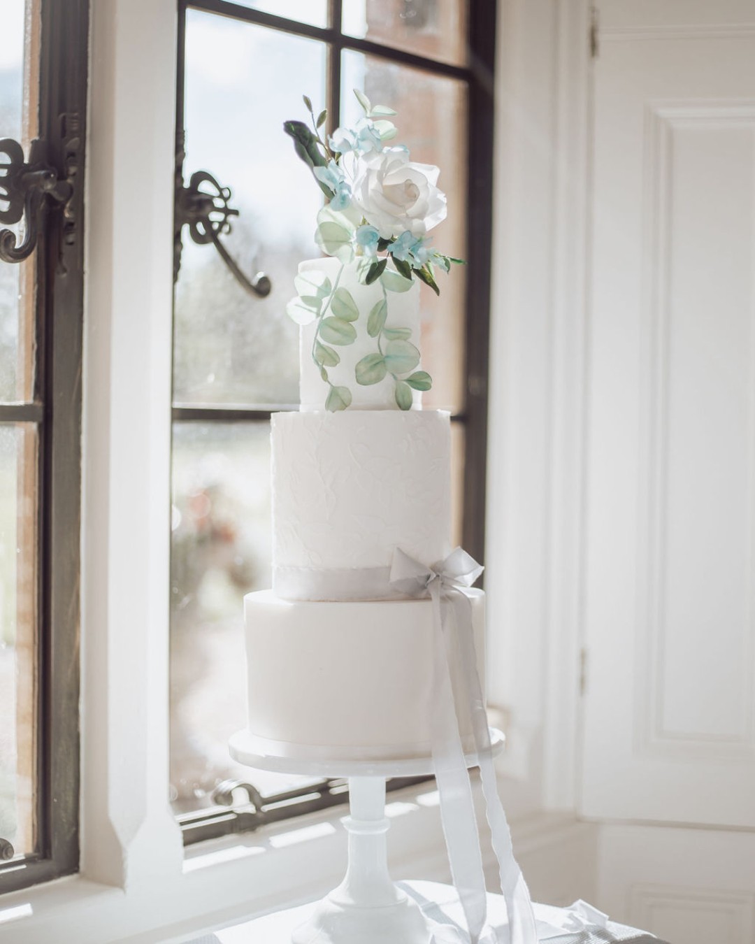 luxury wedding cake surrey. 3 tier cake soft and elegant with lace texture and a satin grey bow. decorated with white and blue sugar florals